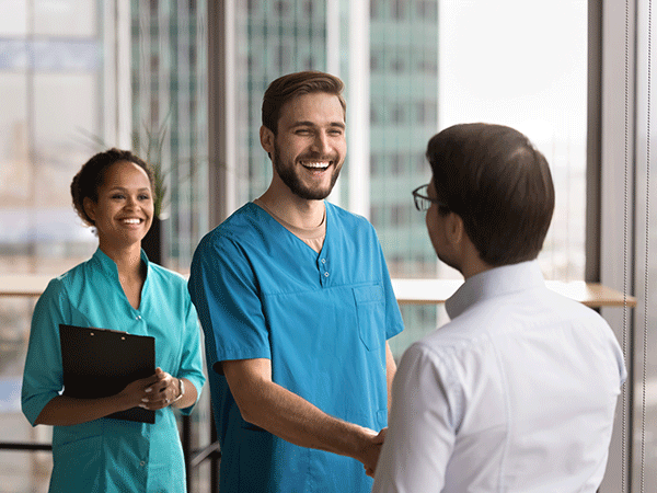 A healthcare professional shaking hands with a business professional. Another healthcare professional is standing in the background.